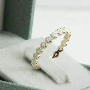Puede incluir: Un delicado anillo de banda de oro con una fila de piedras de &oacute;palo blanco. El anillo se muestra en una caja de anillo de terciopelo verde.