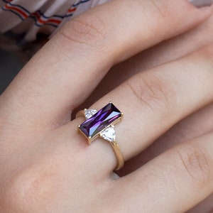May include: A gold ring with a purple rectangular gemstone and two smaller clear heart-shaped gemstones on either side.