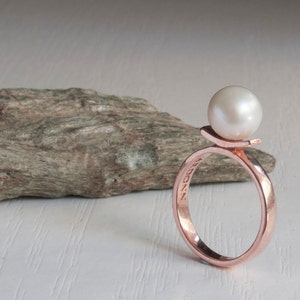 May include: A rose gold ring with a single white pearl set on a raised band. The ring is engraved with the word "BROOKLYN".