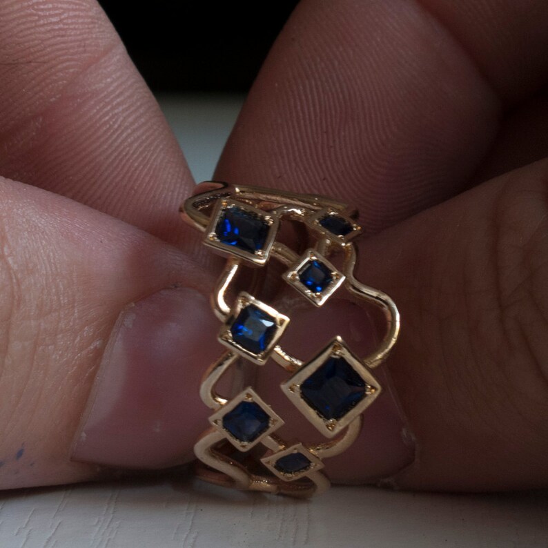 May include: A gold-colored ring featuring a unique design with multiple square and diamond-shaped blue gemstones. The ring's band has an open, flowing design, with the gemstones set at various points along the band.