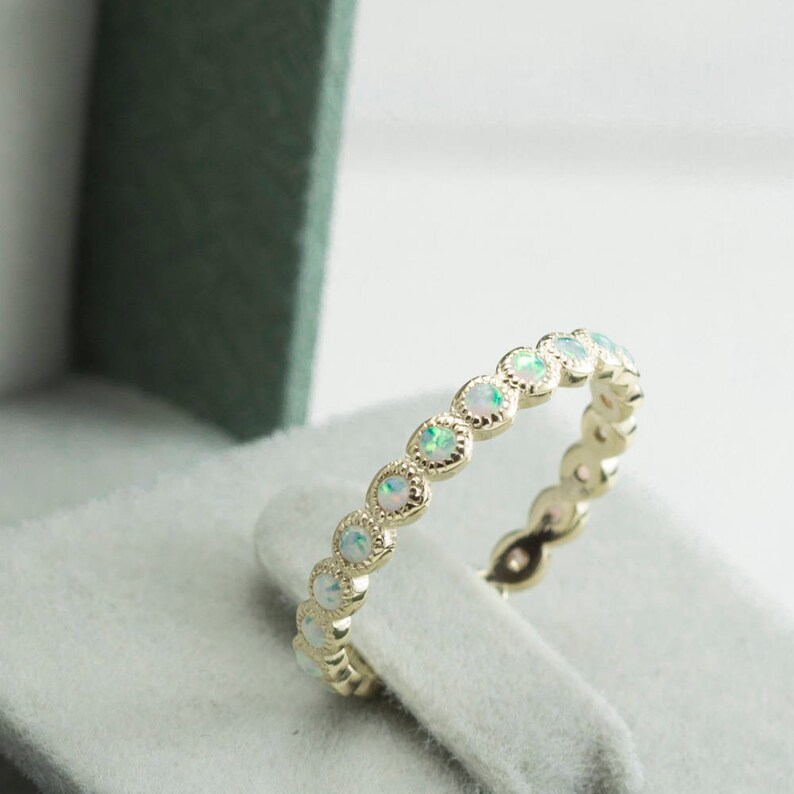 Puede incluir: Un delicado anillo de banda de oro con una fila de peque&ntilde;as piedras de &oacute;palo blanco engastadas en un dise&ntilde;o festoneado.