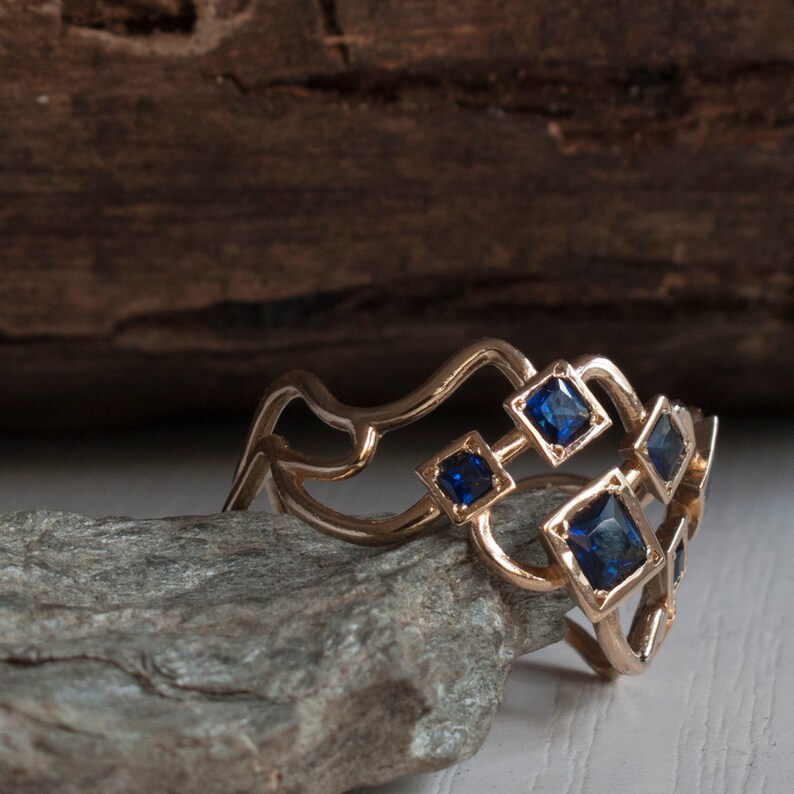 May include: A gold-colored ring with a unique design featuring square, dark blue gemstones. The ring has an open, wave-like structure with the gemstones set in a geometric pattern. The ring is displayed on a gray stone.