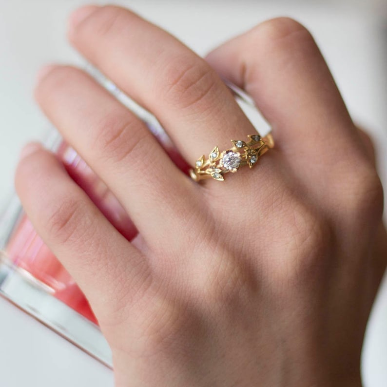 May include: A delicate gold ring with a single round diamond surrounded by a leafy design. The ring is set on a finger.