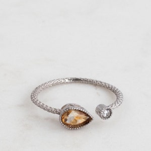 May include: A silver ring with a teardrop-shaped topaz gemstone and a small round diamond accent. The ring is open on one side and has a textured band.