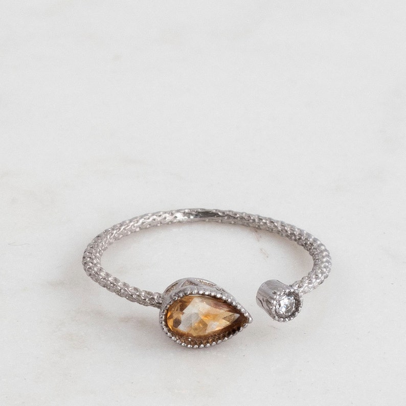 May include: A silver ring with a teardrop-shaped topaz gemstone and a small round diamond accent. The ring is open on one side and has a textured band.