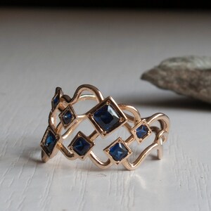 May include: A rose gold ring featuring a geometric design with square, deep blue gemstones. The ring's openwork design incorporates diamond shapes, creating a unique and elegant piece of jewelry.