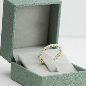 Puede incluir: Un delicado anillo de banda de oro con una fila de peque&ntilde;as piedras de &oacute;palo blanco engastadas en un dise&ntilde;o festoneado. El anillo se muestra en una caja de anillo de terciopelo verde.