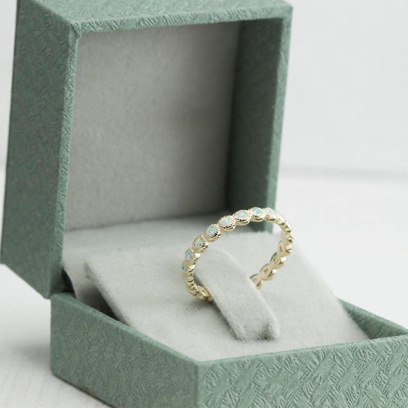 Puede incluir: Un delicado anillo de banda de oro con una fila de peque&ntilde;as piedras de &oacute;palo blanco engastadas en un dise&ntilde;o festoneado. El anillo se muestra en una caja de anillo de terciopelo verde.