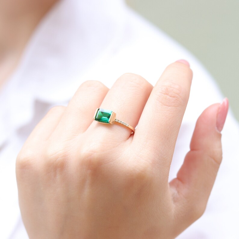 May include: A gold ring with a rectangular emerald green gemstone and small clear gemstones on the band.