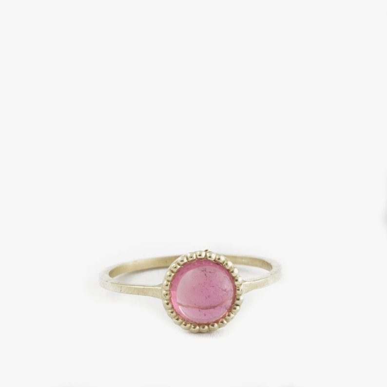 Puede incluir: Un anillo de oro con una piedra de vidrio rosa engastada en un bisel de cuentas.