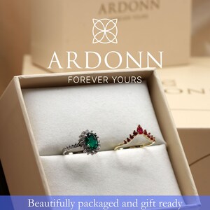 A white jewelry box containing two rings, one with a green gemstone and the other with a red gemstone, with the Arddon logo and text above the box.