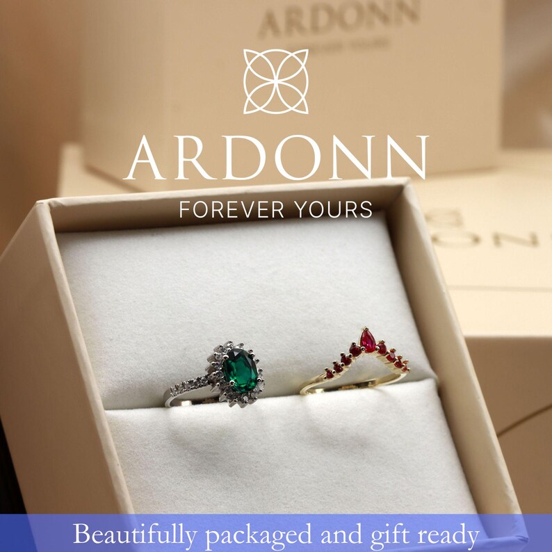 A white jewelry box containing two rings, one with a green gemstone and the other with a red gemstone, with the Arddon logo and text above the box.