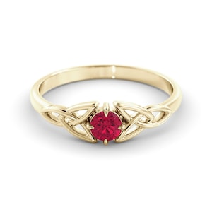 May include: A gold-colored ring featuring a central, round, red gemstone. The ring's band has a Celtic knot design on either side of the gemstone, adding a touch of intricate detail. The ring is set against a white background.