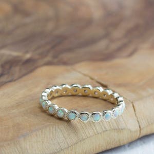 Puede incluir: Un delicado anillo de banda de oro con una fila de peque&ntilde;as piedras redondas de &oacute;palo blanco.