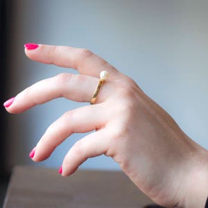May include: A gold ring with a single white pearl on the top. The ring is worn on a finger with pink nail polish.