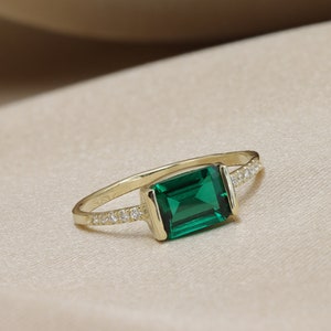 May include: A gold ring with a large emerald green gemstone and a row of small clear gemstones on the band.