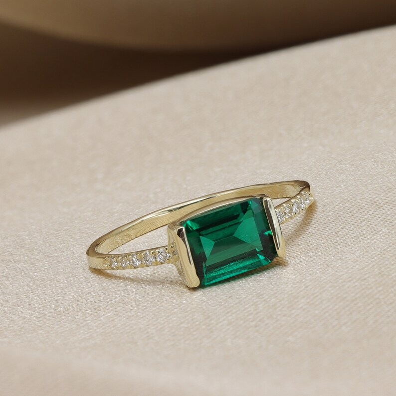 May include: A gold ring with a large emerald green gemstone and a row of small clear gemstones on the band.