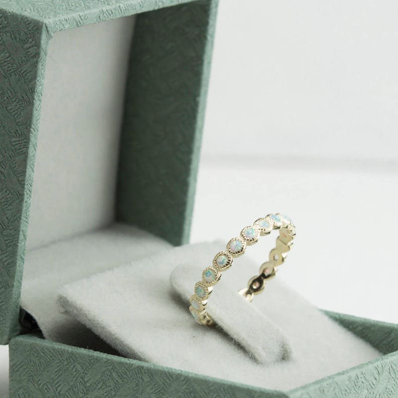 Puede incluir: Un delicado anillo de banda de oro con una fila de piedras de &oacute;palo blanco. El anillo se muestra en una caja de anillo de terciopelo verde.
