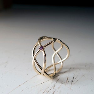 May include: A gold ring with a delicate, interwoven design and a small, dark red gemstone in the center.