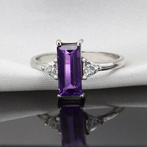 May include: A silver ring with a rectangular purple amethyst gemstone and two smaller triangular clear gemstones on either side of the amethyst.