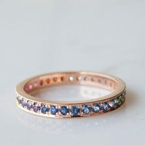 May include: A rose gold ring with a band of colorful gemstones. The ring features a continuous row of various colored stones, including pink, orange, blue, green, and purple. The ring is set against a white background.