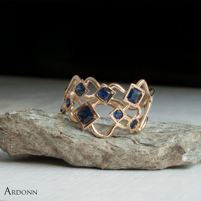 May include: A gold-colored ring with a unique openwork design. The ring features multiple square, dark blue gemstones set within the intricate metalwork. The ring is displayed on a gray stone surface. The brand name "ARDONN" is visible in the lower left corner.