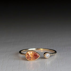 Puede incluir: Un anillo de oro con una piedra preciosa naranja en forma de lágrima y una pequeña piedra preciosa redonda transparente. El anillo está abierto por un lado.