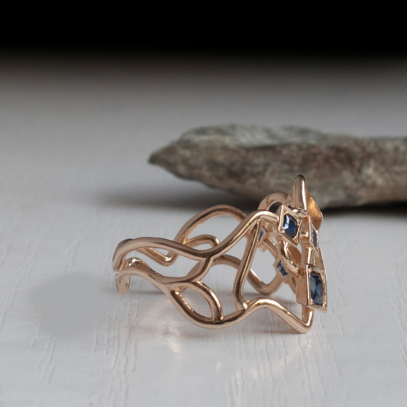 May include: Rose gold ring with a unique, openwork design. The ring features a series of flowing lines and geometric shapes, with several small, square, dark blue gemstones set into the design. The ring is displayed on a white surface.