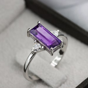 May include: A silver ring with a rectangular purple amethyst gemstone and two smaller triangular clear gemstones on either side of the amethyst.