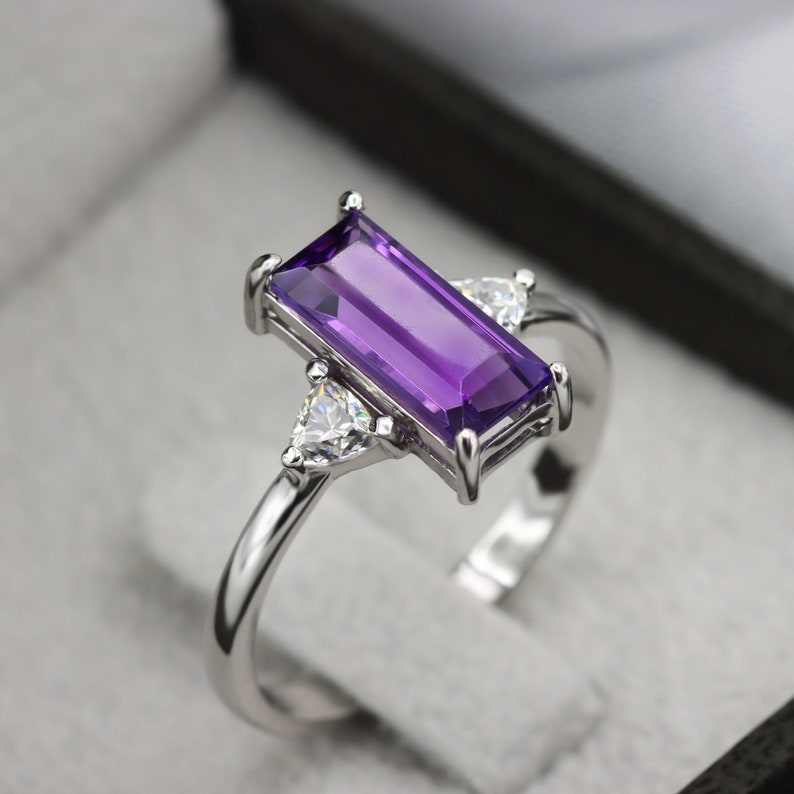 May include: A silver ring with a rectangular purple amethyst gemstone and two smaller triangular clear gemstones on either side of the amethyst.