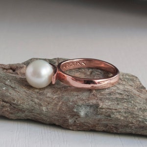 May include: A rose gold ring with a single white pearl. The ring is engraved with the word "ARCONY".