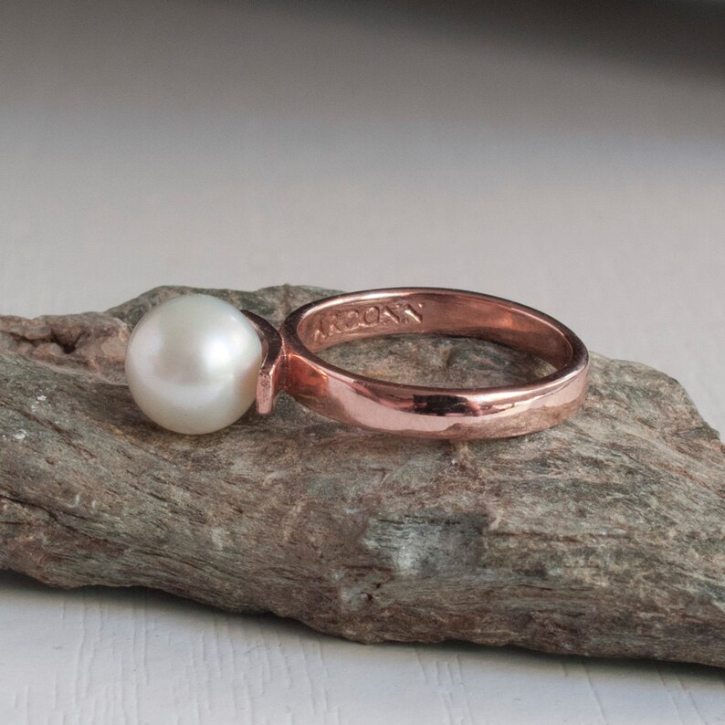 May include: A rose gold ring with a single white pearl. The ring is engraved with the word "ARCONY".