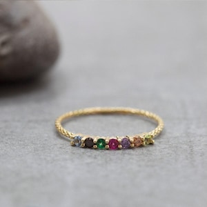 May include: A delicate gold ring featuring a series of colorful gemstones. The ring's band has a textured design, and the gemstones are arranged in a row, showcasing a spectrum of colors. The ring is set against a neutral background.