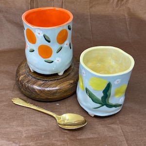 May include: Two handmade ceramic cups with irregular shapes and small feet. One cup features orange fruit and white flower patterns with a bright orange interior. The other cup displays yellow lemon patterns with green leaves and a yellow interior. A gold spoon is included.