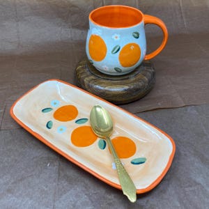 May include: A ceramic mug with a light blue exterior and orange interior, featuring painted orange fruit and small white flowers. A matching rectangular ceramic tray with orange borders and orange fruit motifs, and a gold-toned metal spoon.