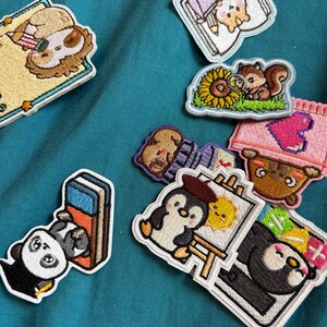 Puede incluir: Una colección de parches bordados con varios personajes, incluyendo un perezoso, un hámster con un lápiz, una ardilla con un girasol, un panda con una goma de borrar, un pingüino pintando un sol y un oso asomándose tras un corazón rosa.