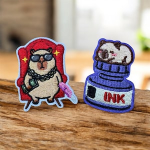 May include: Two embroidered patches. The left patch features a tan capybara wearing sunglasses and a chain, sitting in a red chair while holding money. The right patch shows a small creature resting on a blue ink bottle with the text INK printed on it.