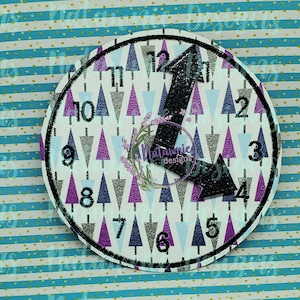 May include: A white clock face with black numbers and a black hand pointing to the number 12. The clock face is surrounded by a black circle and has a pattern of purple, blue, and white triangles. The text "Matannie Designs" is visible in the center of the clock face.