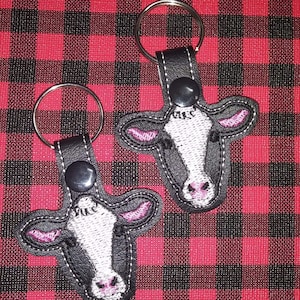 May include: Two black leather keychains with embroidered cow faces. The cow faces are white with black spots and have pink noses and ears.
