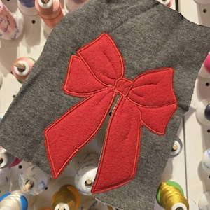 May include: A gray fabric with a red felt bow appliquéd on it. The fabric is surrounded by spools of thread.