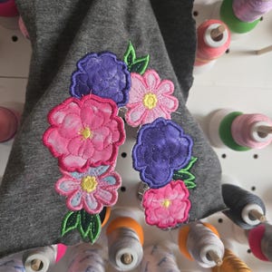 May include: A gray fabric item with embroidered pink, purple, and light blue flowers and green leaves. The flowers have yellow centers. The background includes spools of thread in various colors.