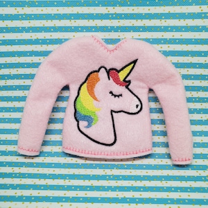 May include: Pink fleece sweater with a rainbow unicorn applique. The unicorn has a closed eye and a gold horn.