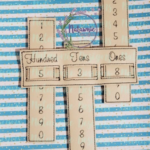 May include: A beige felt board with three columns labeled "Hundreds", "Tens", and "Ones". Each column has a set of numbers from 0 to 9. The numbers are arranged vertically in each column. The board is on a blue and white striped background.