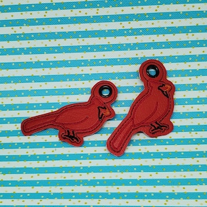 May include: Two red felt cardinal bird ornaments with black eyes and beaks. The ornaments have a hole at the top for hanging. The ornaments are on a blue and white striped background.