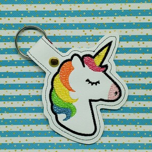 May include: White leather keychain with a rainbow unicorn embroidered on it. The unicorn has a closed eye and a pink horn.