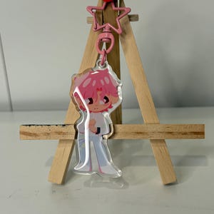 May include: A clear acrylic keychain featuring a cartoon character with pink hair and a white shirt. The keychain is attached to a pink star-shaped charm and a red push pin. The keychain is displayed on a small wooden easel.