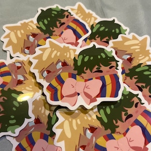May include: A stack of colorful stickers featuring two cartoon characters hugging. The characters are wearing striped shirts with a pink bow. The characters have green and yellow hair.