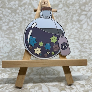 Peut inclure: Une bouteille en verre violette avec un bouchon en liège remplie d'un liquide foncé et une étiquette avec deux yeux. La bouteille est décorée d'étoiles colorées.