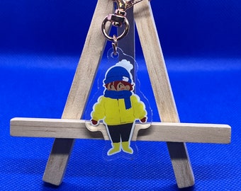 May include: A clear acrylic keychain featuring a cartoon character wearing a yellow jacket, blue hat, and scarf. The keychain is attached to a gold-colored metal clasp with a rainbow heart charm.