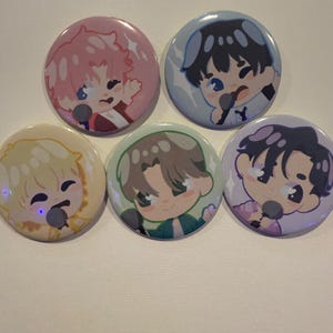 May include: Five round pinback buttons with cartoon illustrations of people singing into microphones. The buttons have different colored backgrounds, including pink, blue, yellow, green, and purple. The illustrations have a cute, anime-style design.